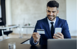 Man in blue suit with black tie. Holding a white phone and card smiling infront of a laptop. Making a purchase.