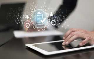 hand using a tablet on a dark wooden table. Graphic above the hand showing a clipboard with a tick through it