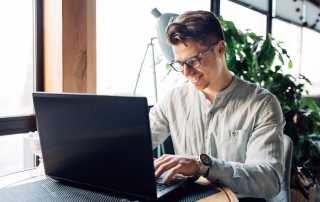 man with glasses on smiling as he works on a laptop in a brightly lit room.