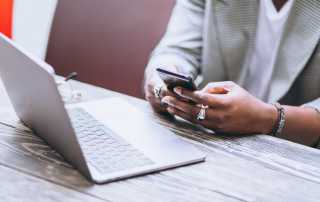 person sat on a wooden table with an open laptop. Person is looking at their smartphone.