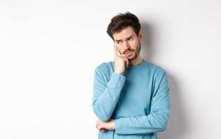 Worried young man with beard in sweatshirt, looking away pensive and thinking, standing troubled against white background.