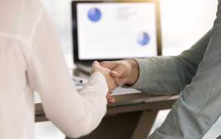 Close-up of a handshake between two professionals, with a laptop displaying charts in the background.