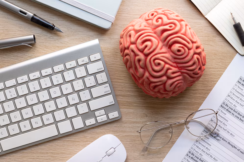 Artificial brain model on a wooden desk surrounded by a keyboard, glasses and documents.